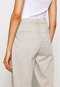 Světlé béžové kalhoty vyrobené z měkké tkaniny, s uvolněným střihem, dvěma zadními kapsami s jemným švem a hladkou texturou.