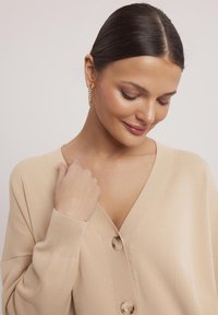 Beżowy kardigan z dekoltem w kształcie litery V, ściągaczowymi mankietami i trzema dużymi guzikami. Miękka tekstura i luźny krój, charakteryzujący się minimalistycznym designem.