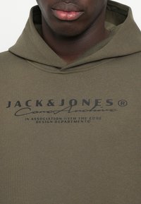 Hoodie vert olive en mélange de coton, avec un logo imprimé en noir sur le devant. Le tissu présente une finition lisse et texturée.
