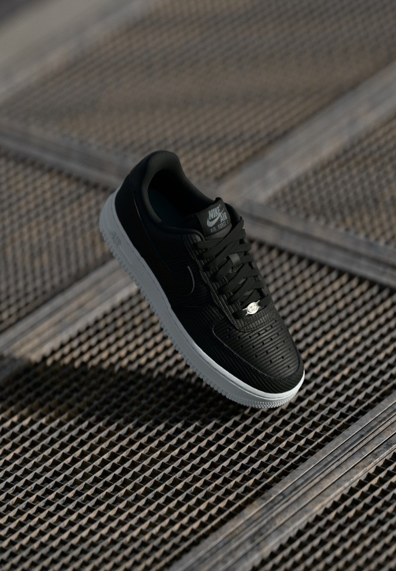 Crne Nike Air Force 1 tenisice s teksturiranim gornjim dijelom, ravnim crnim vezicama i bijelim međuslojem. Sadrže logotip na jeziku i bočnoj strani.