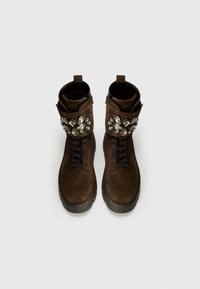 Stivaletti alla caviglia in suede marrone con un design con lacci, arricchiti da grandi dettagli metallici sulla caviglia, con suola nera e dettagli con zip.