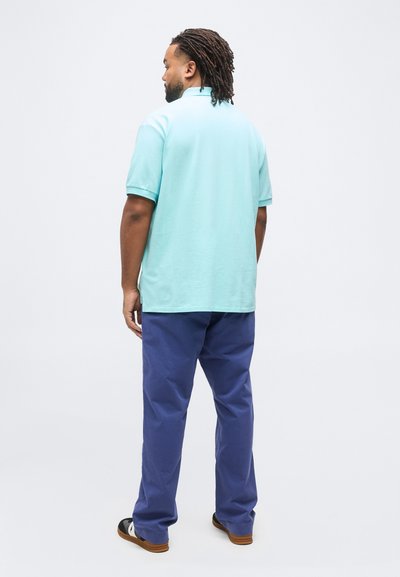 Άνδρας με ράστα, όρθιος με την πλάτη στραμμένη, φοράει ανοιχτό μπλε πόλο μπλούζα, μπλε παντελόνι και μαύρα και άσπρα παπούτσια, μπροστά σε απλό φόντο.