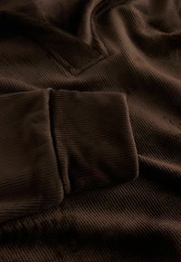 Hnědá kordová tkanina s ribbovanou strukturou, s ohrnutým manžetovým lemem a mírným výstřihem do V, vytvářející jemný, teplý vzhled.