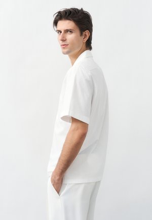 Νέος άνδρας με σκούρα μαλλιά, φοράει λευκό κοντομάνικο πουκάμισο και λευκό παντελόνι, στέκεται πλευρικά με τα χέρια στις τσέπες.