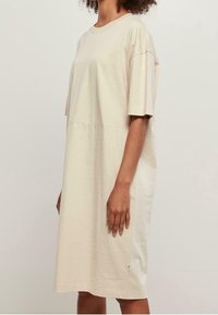 Beige, ylivaltainen T-paitamekko, joka on valmistettu pehmeästä puuvillasta. Lyhyet hihat, pyöreä pääntie ja sivuhalkio, jossa pieni yksityiskohta helmassa.