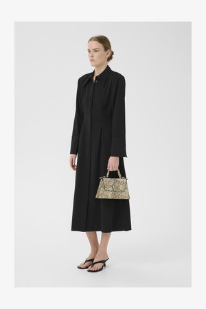 Vestido preto de mangas compridas com colarinho, design em A. Leva uma pequena mala em padrões de pele de serpente em bege e marrom. Chinelos pretos.