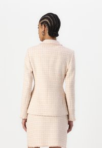 Blazer de tweed ajustado em rosa claro com um acabamento texturizado, apresentando um colarinho em ângulo e uma silhueta ajustada, com uma saia a condizer abaixo.