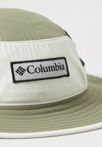 Columbia šešir u obliku kante u maslinasto zelenoj i bijeloj boji, izrađen od tkanine s glatkom teksturom; sadrži istaknuti crni logotip.