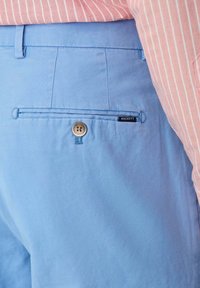 Pantalons bleu clair en coton avec une poche arrière, comportant un bouton et une petite étiquette noire avec le nom de la marque "HACKETT."