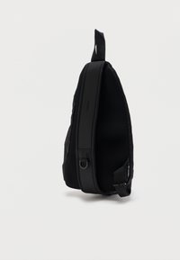 Μαύρο σακίδιο ώμου με επενδυμένη πλέξη στην πλάτη, ρυθμιζόμενο ιμάντα, λαβή βρόχου και μικρή ετικέτα με το λογότυπο σε λευκό φόντο.