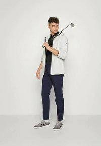 Pánske športové oblečenie, ktoré obsahuje svetlošedú bundu na zips, čierne polo tričko, námornícke nohavice a šedé tenisky, držiace golfovú palicu.