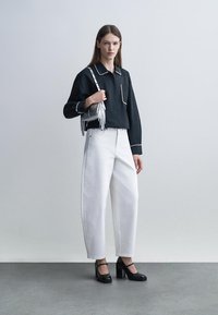 Moteris apsirengusi juodą ilgomis rankovėmis marškinėlį su balta apdaila, baltas plataus kirpimo kelnes, juodus aukštakulnius ir nešiojanti sidabrinę rankinę su kutais.