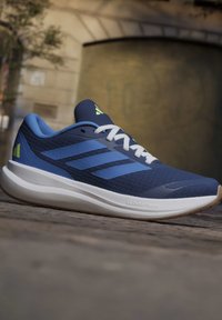 Μπλε παπούτσι τρεξίματος Adidas με λευκά κορδόνια και απορροφητική λευκή σόλα με την ένδειξη Cloudfoam, τοποθετημένο σε εξωτερική πλακόστρωση.