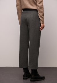 Grijze, gestructureerde broek met een losse pasvorm en een zwarte elastische tailleband, gecombineerd met zwarte enkellaarzen. Gezicht vanuit de achterkant.