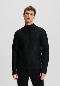 Sort strikket sweater med en høj ribbet krave, prydet med et struktureret diamantmønster. Ribbet manchetter og kant giver et tætsiddende udseende.