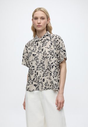 Woman with blonde hair wearing a beige short-sleeve blouse with black floral pattern and white wide-leg pants, standing with neutral expression.