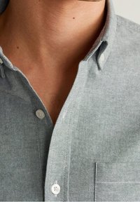 Camisa de manga corta con cuello en tejido ligero gris claro con un acabado texturizado. Presenta un bolsillo en el pecho y botones blancos.
