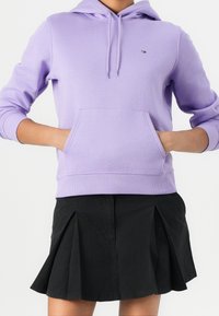 Sudadera con capucha color lila con bolsillo tipo canguro, puños y dobladillo acanalados. Presenta un pequeño logo en el pecho. Combinada con una falda plisada negra.