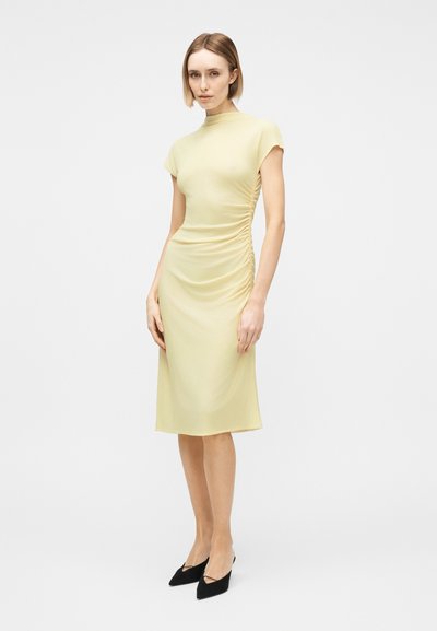 Γυναίκα με κοντά ξανθά μαλλιά που φοράει εφαρμοστό παλ ανοιχτό κίτρινο μίντι φόρεμα με πιέτες στο πλευρό και μαύρες μυτερές γόβες, στέκεται μπροστά σε λευκό φόντο.