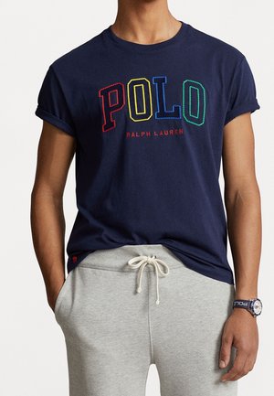 Osoba nosząca granatowy t-shirt Polo Ralph Lauren z wielokolorowymi literami oraz jasnoszare spodnie dresowe ze sznurkiem.
