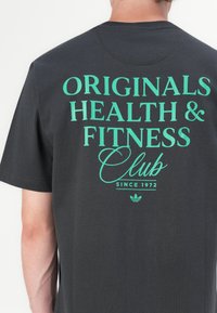 T-shirt en coton noir avec un grand texte vert au dos : "ORIGINALS HEALTH & FITNESS Club SINCE 1972" avec le logo Adidas en dessous.