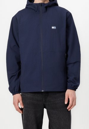 Mand iført navy hættetrøje med lynlås og lille Tommy Jeans-logo på brystet og sorte jeans, står mod hvid baggrund.