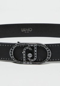 Cintura in pelle nera con una fibbia decorativa adornata di strass e un logo, arricchita da borchie metalliche lungo la cintura.