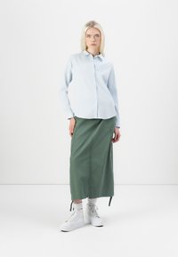 Hellblaues Hemd mit Kragen, kombiniert mit einem langen grünen Rock mit Seitentaschen. Weiße Plateausneaker runden das Outfit ab.