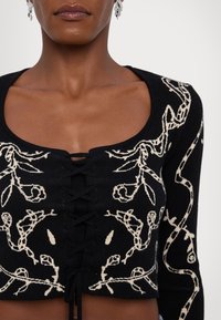 Haut court noir à motif de vigne blanche avec un laçage à l'avant et un décolleté carré, porté par une personne à peau foncée.