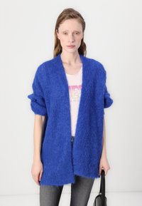 Blå, fluffy cardigan med løs passform og rullesleeves, åpen frontdesign, kombinert med en lys rosa topp og mørke jeans.