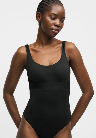 Γυναίκα με πλεξούδες που φοράει εφαρμοστό μαύρο ολόσωμο μαγιό χωρίς μανίκια, στέκεται με τα χέρια ελαφρώς πίσω από την πλάτη της.