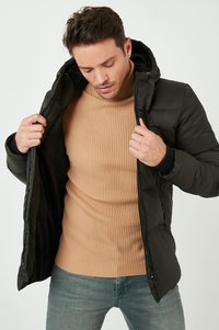 Giacca a vento nera con cappuccio, con un'esterno liscio e chiusura a zip, indossata sopra un maglione a collo alto a coste marrone chiaro.