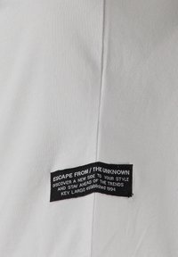 Etiqueta de tela negra cosida en una prenda blanca con el texto "Escapa de lo desconocido, descubre un nuevo lado de tu estilo, Key Largo fundado en 1994."