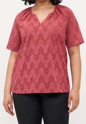 Vrouw draagt een kort mouwig stofroze blouse met kantpatroon en V-hals, gecombineerd met zwarte nauwsluitende broek.