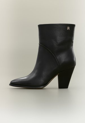 Bottine en cuir noir avec bout pointu, talon bloc et petit logo métallique près du sommet sur fond neutre.