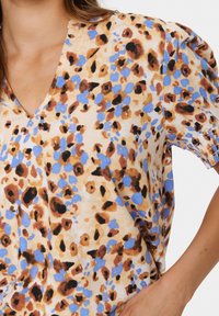 T-shirt à manches courtes en tissu léger avec un imprimé floral abstrait beige, bleu et marron et un décolleté en V. Détails plissés sur les manches.