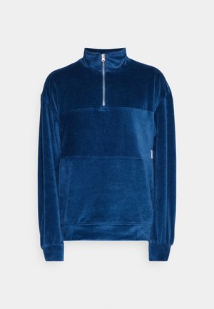 Granatowy welurowy sweter z długimi rękawami, wysokim kołnierzem i krótkim srebrnym zamkiem z przodu na jasnoszarym tle.