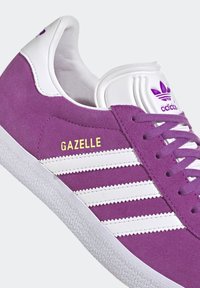 Baskets en suede violet avec des accents en cuir blanc et trois rayures horizontales. "GAZELLE" en texte doré ; semelle en caoutchouc texturé blanc.