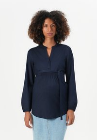 Blusa premaman blu navy con maniche lunghe, abbottonatura frontale, dettaglio in vita arricciato e un fiocco. Tessuto morbido con colletto strutturato e orlo rotondo.