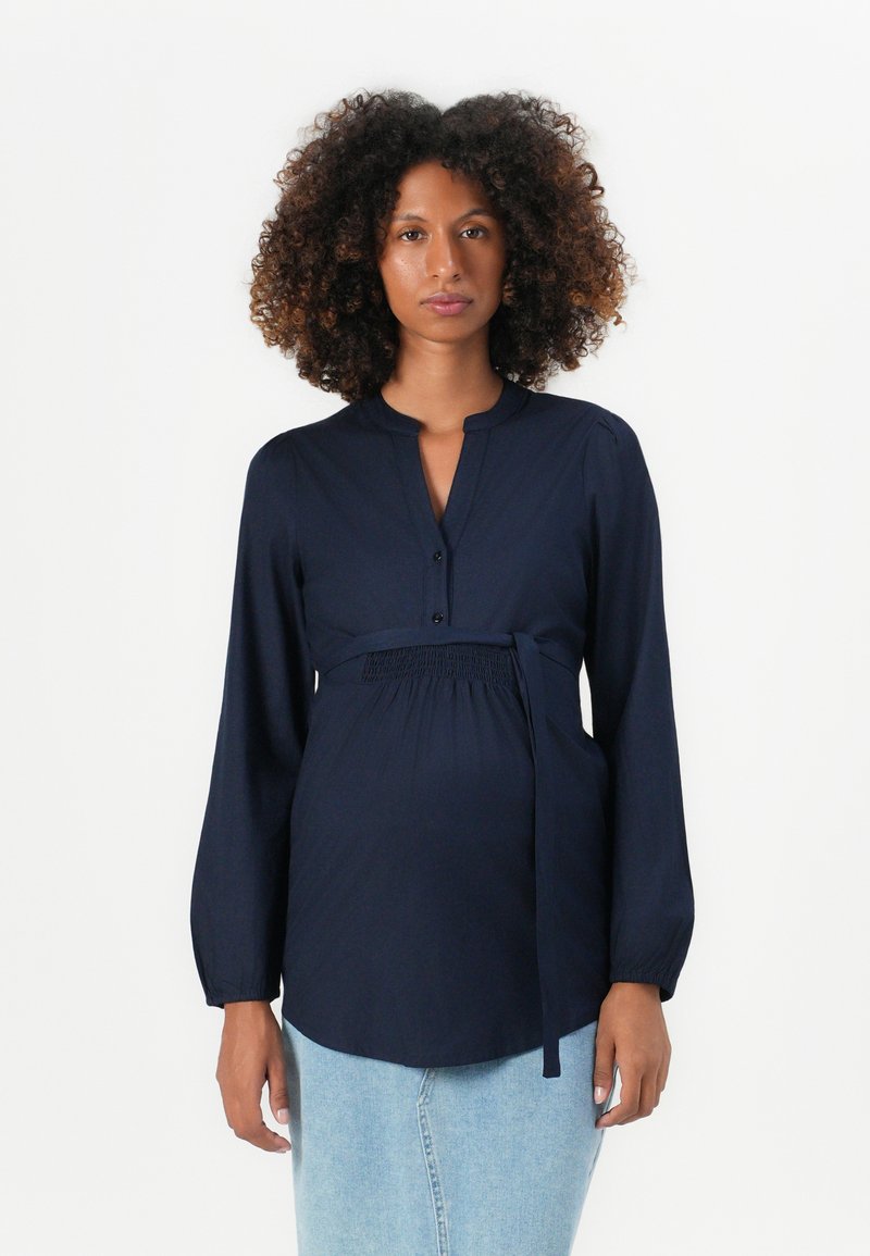 Blusa premaman blu navy con maniche lunghe, abbottonatura frontale, dettaglio in vita arricciato e un fiocco. Tessuto morbido con colletto strutturato e orlo rotondo.