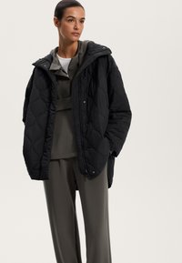 Sort quiltet jakke med hætte, frontknaplukning og en afslappet pasform, lagd over en grå hættetrøje og matchende bukser.