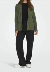 Zielony żakiet z dwoma guzikami i przednimi kieszeniami, w połączeniu z czarnymi spodniami z szerokimi nogawkami i białymi sneakersami, na jednolitym tle.