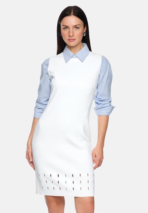 Woman wearing a white sleeveless dress with a perforated hem over a blue and white striped shirt with rolled-up sleeves.