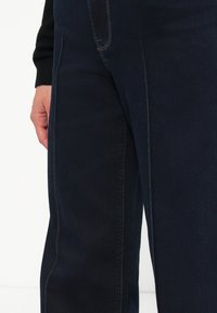 Pantalones de mezclilla azul oscuro con una textura suave, que presentan costuras en contraste y un diseño ajustado, con una mano visible cerca de la cintura.