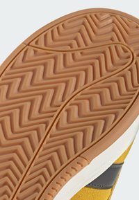 Semelle de chaussure en caoutchouc avec un motif de bande de roulement en zigzag, de couleur beige et surface texturée. Accentuée par un bord blanc et une section supérieure jaune.