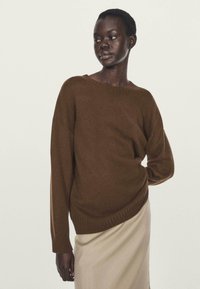 Donna con i capelli corti che indossa un maglione di maglia marrone e una gonna beige, in piedi con una mano sulla schiena contro uno sfondo neutro.