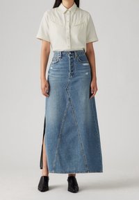 Denim maxikjol med en fram slit, har en åtsittande midja, ljusblå färg och en distinkt vinklad sömdesign. Bärs med en ljus skjorta.