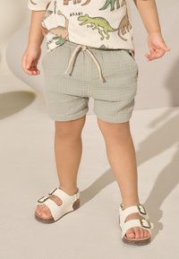 Pantaloncini verdi con texture e vita elasticizzata, abbinati a sandali bianchi con due fibbie dorate e suola marrone.