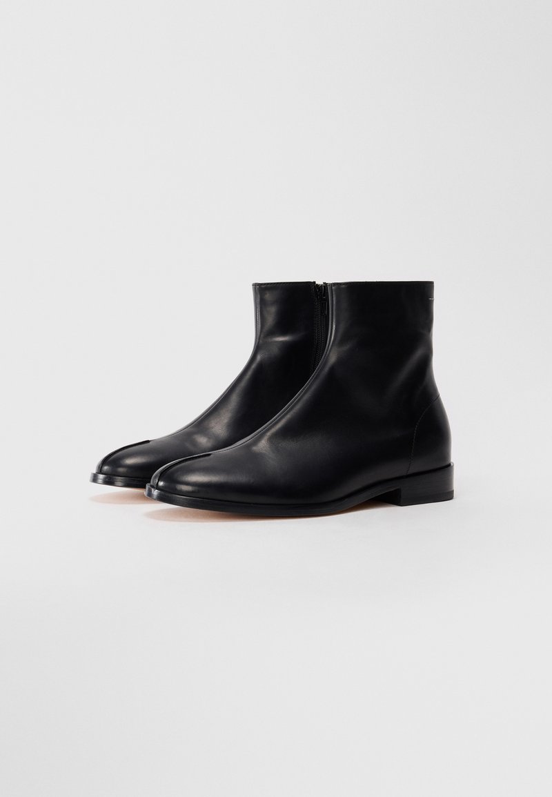 Une paire de bottes noires en cuir à la cheville avec des fermetures éclair latérales et un design à bout fendu, présentée sur un fond blanc uni.