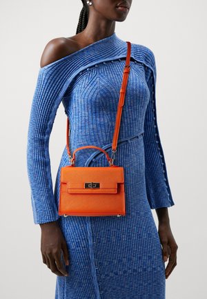 Sac à main texturé orange avec une poignée supérieure et une bandoulière réglable, doté d'une fermeture métallique au centre. Porté avec une robe côtelée bleue.